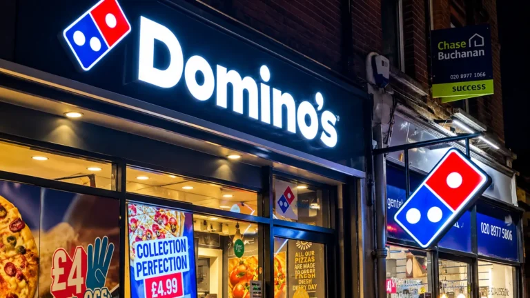 Domino’s Pizza storefront at night featuring illuminated brand signage, a hanging logo sign, brightly lit windows with pizza promotions, and a warm interior against a dark urban street.
